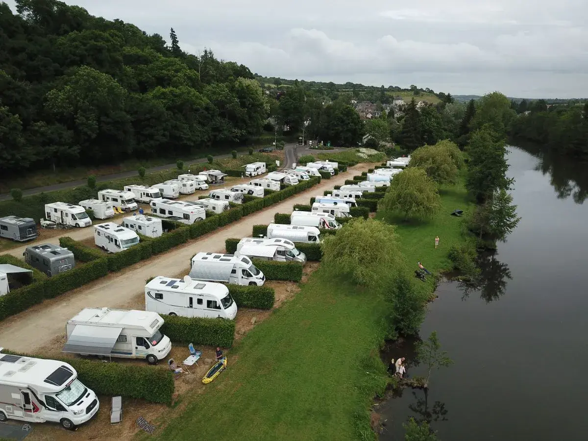 Vue aérienne d'une aire de camping-car gratuite en bord de mer normandie, avec de nombreux véhicules stationnés près d'une rivière bordée d'arbres.