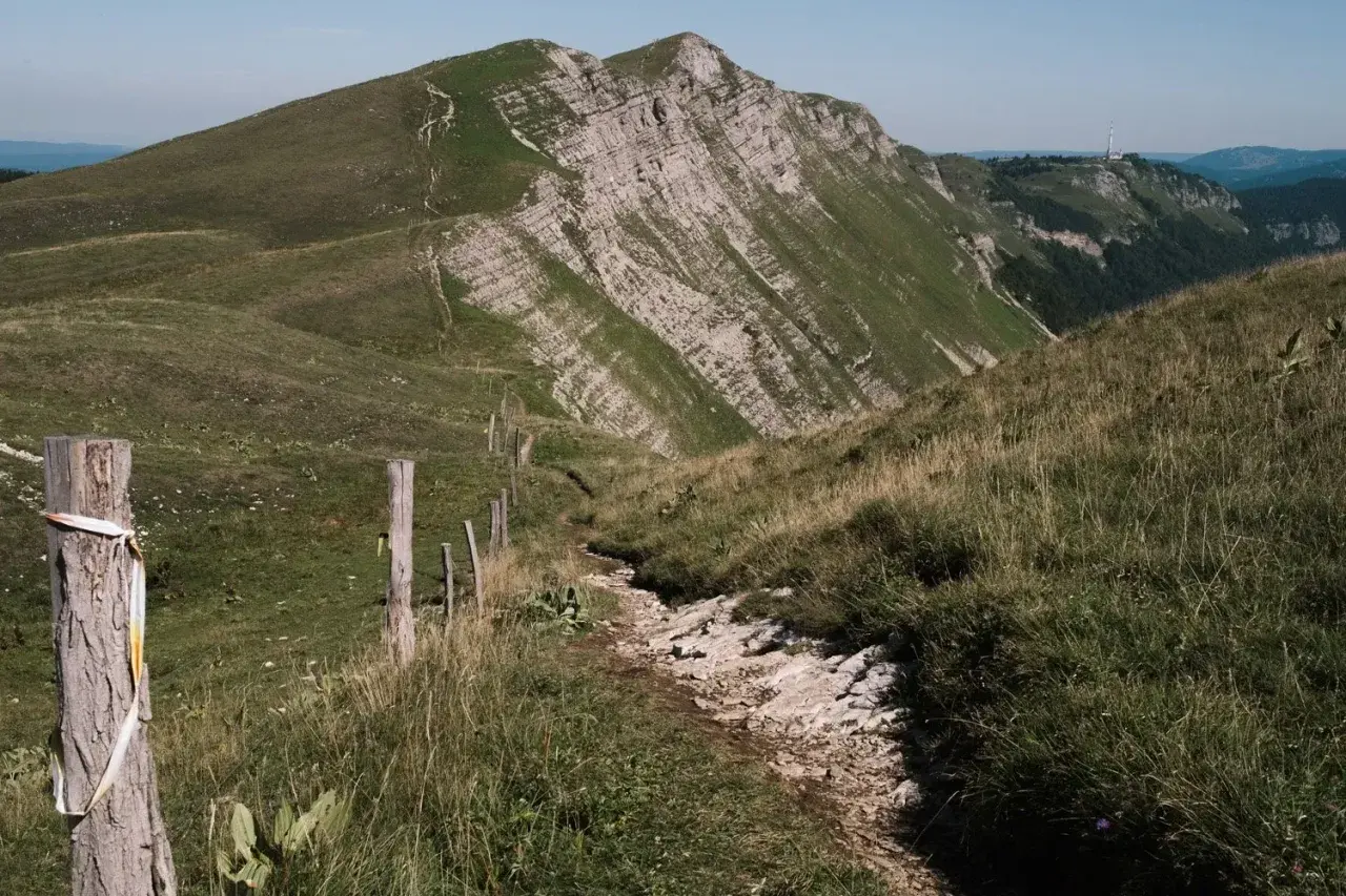 Sentier rocailleux serpentant dans l'herbe haute, menant vers des montagnes escarpées. Une clôture rudimentaire marque le chemin de cette échappée jurassienne.