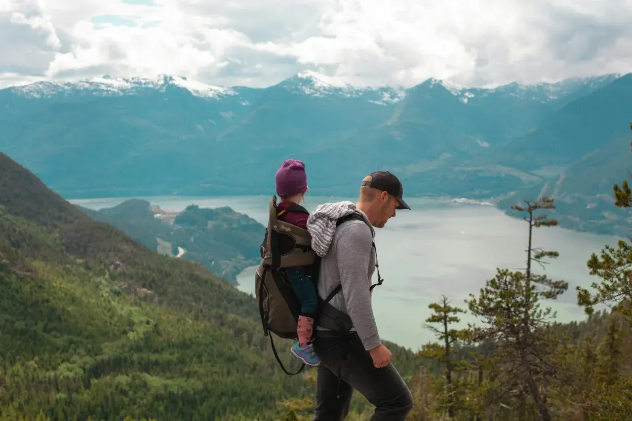 Un père porte son enfant dans un porte-bébé lors d'un week-end randonnée, avec des montagnes enneigées en arrière-plan.