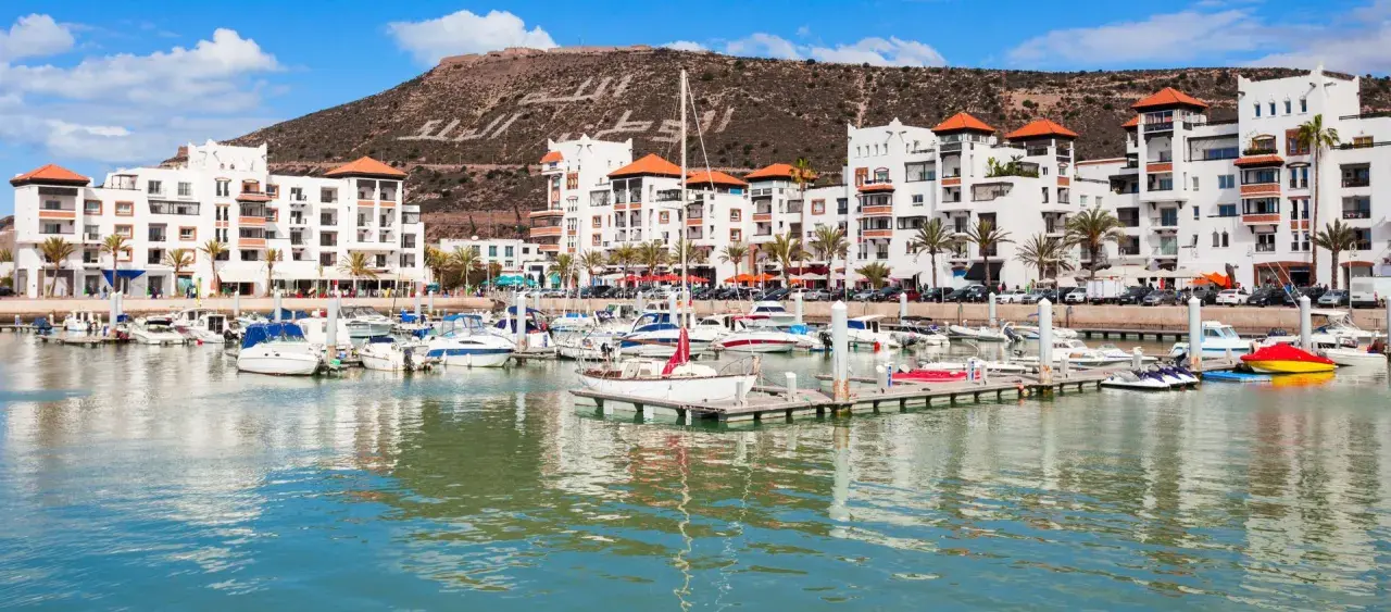 Port de plaisance à Agadir, Maroc, avec des bateaux blancs et des bâtiments modernes. La colline derrière porte une inscription en arabe.