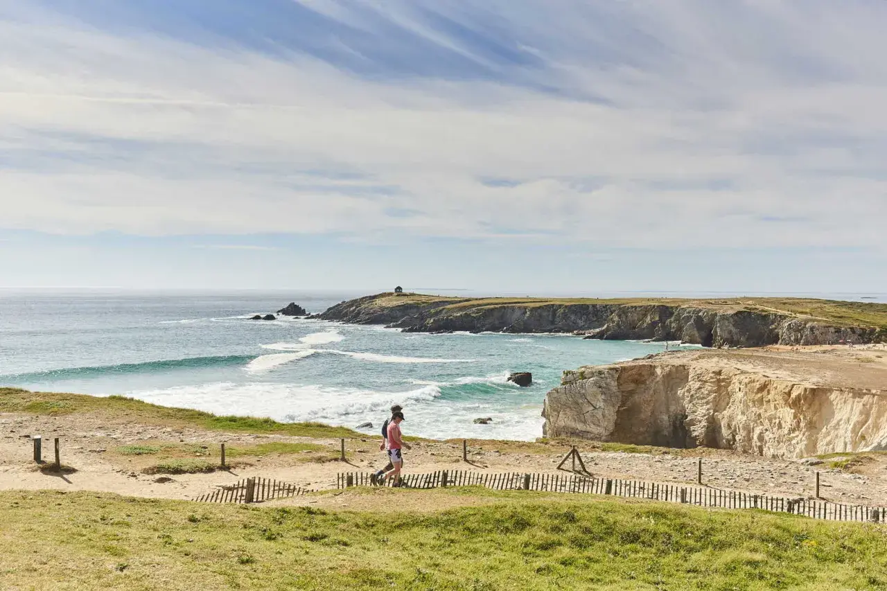 Deux personnes marchent sur un sentier côtier, découvrant les plus beaux endroits de Quiberon. Falaises dorées, mer turquoise et ciel bleu.