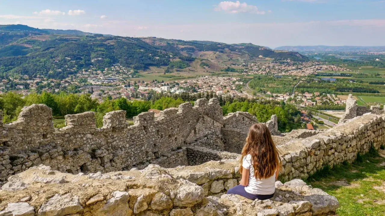 Une jeune fille contemple la vue depuis les ruines du **château de Crussol**, dominant une ville et des collines verdoyantes sous un ciel bleu.