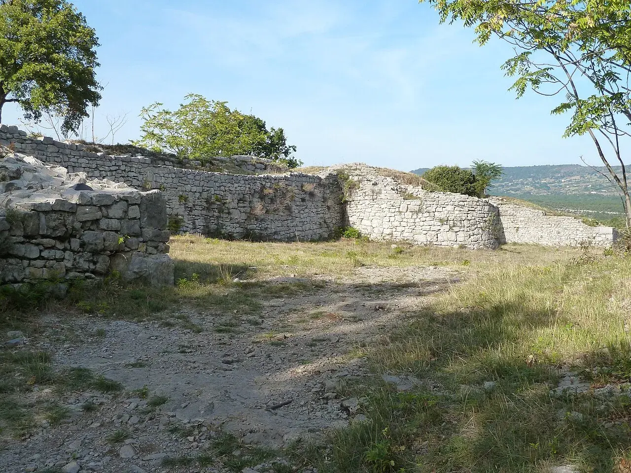 Ruines d'un oppidum gaulois en pierre, avec des murs et des arbres sous un ciel bleu.