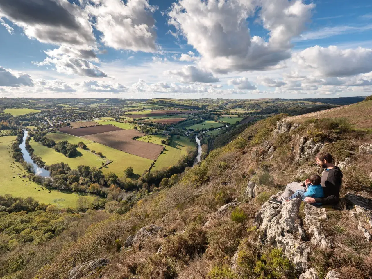Vue panoramique de la Suisse Normande, une carte vivante de collines verdoyantes, d'une rivière sinueuse et d'un père et son enfant contemplant le paysage.