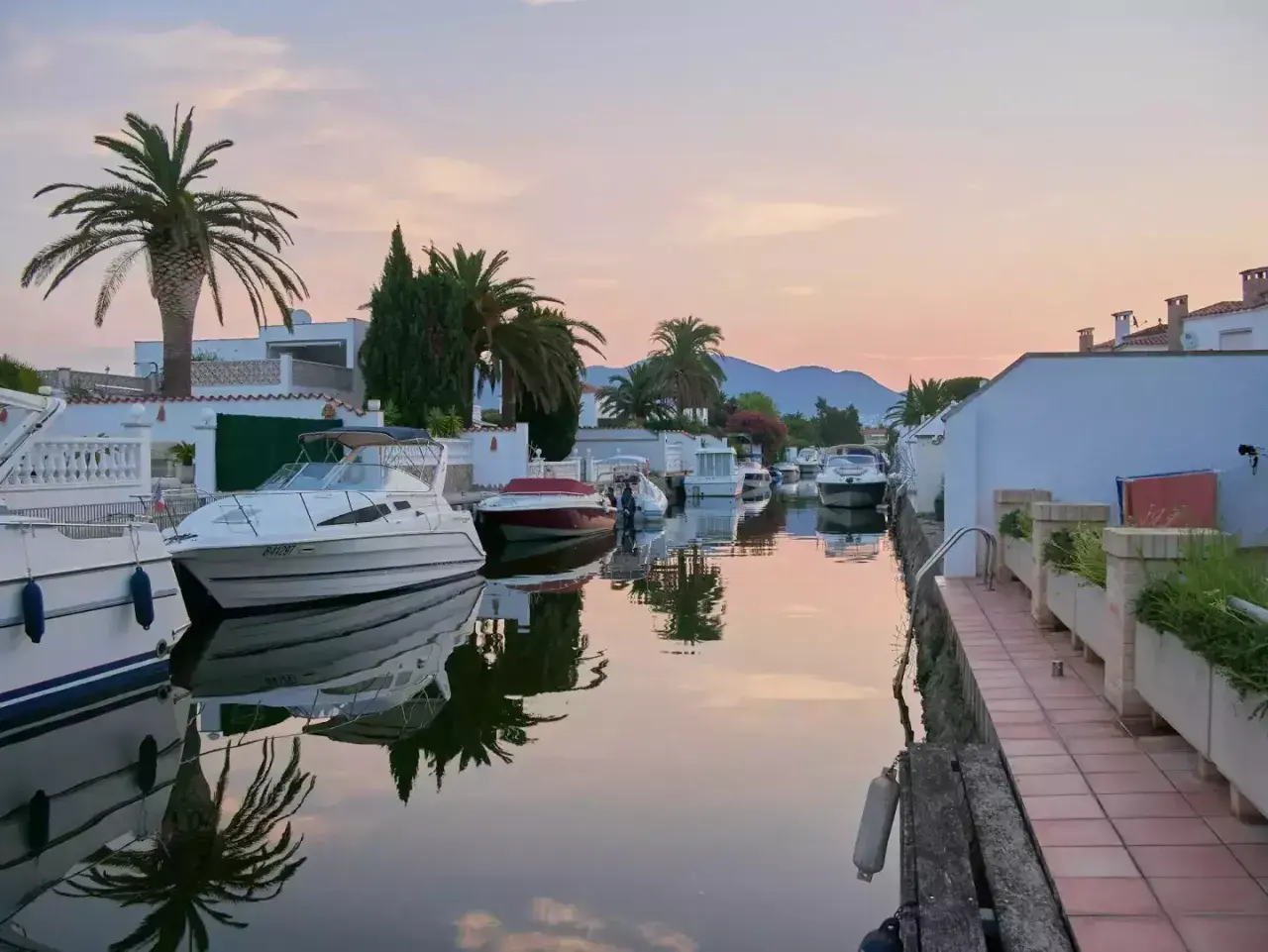 Coucher de soleil paisible sur les canaux d'Empuriabrava, Espagne, avec des bateaux amarrés et des palmiers se reflétant dans l'eau calme.