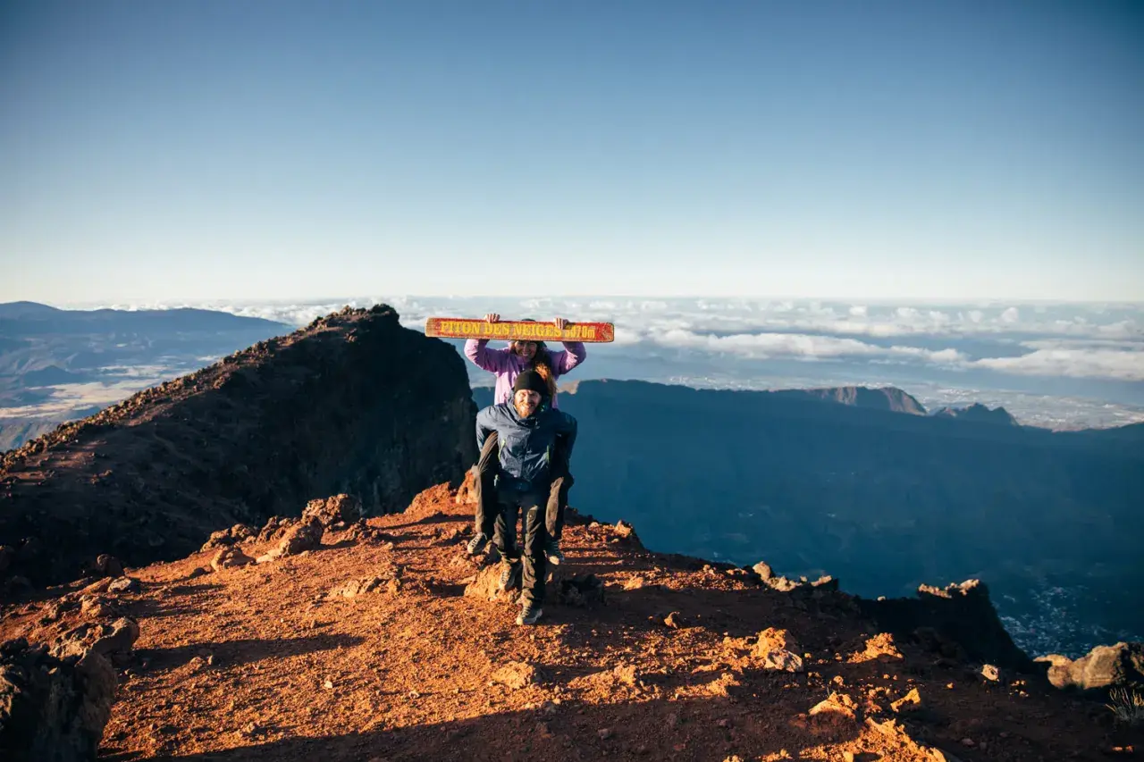 Couple joyeux au sommet du Piton des Neiges, portant une pancarte "Piton des Neiges 3070m". Paysage spectaculaire au lever du soleil.