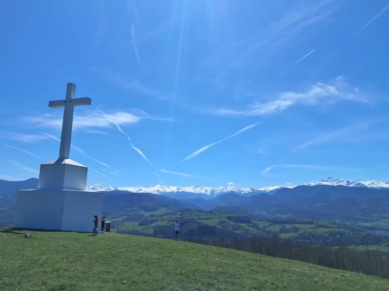 Le tuc de Montcalivert domine un paysage verdoyant sous un ciel bleu zébré de traînées de condensation. Une grande croix blanche se dresse fièrement.