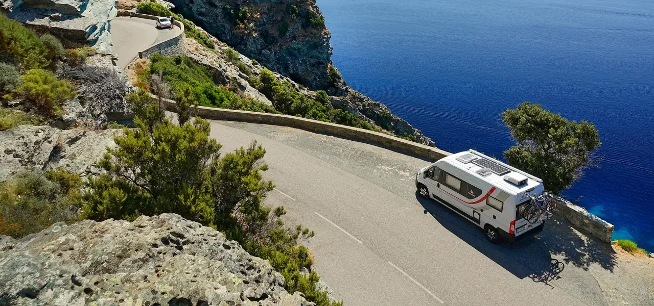 Un camping-car blanc circule sur une route côtière sinueuse en Corse, dominant la mer d'un bleu profond.