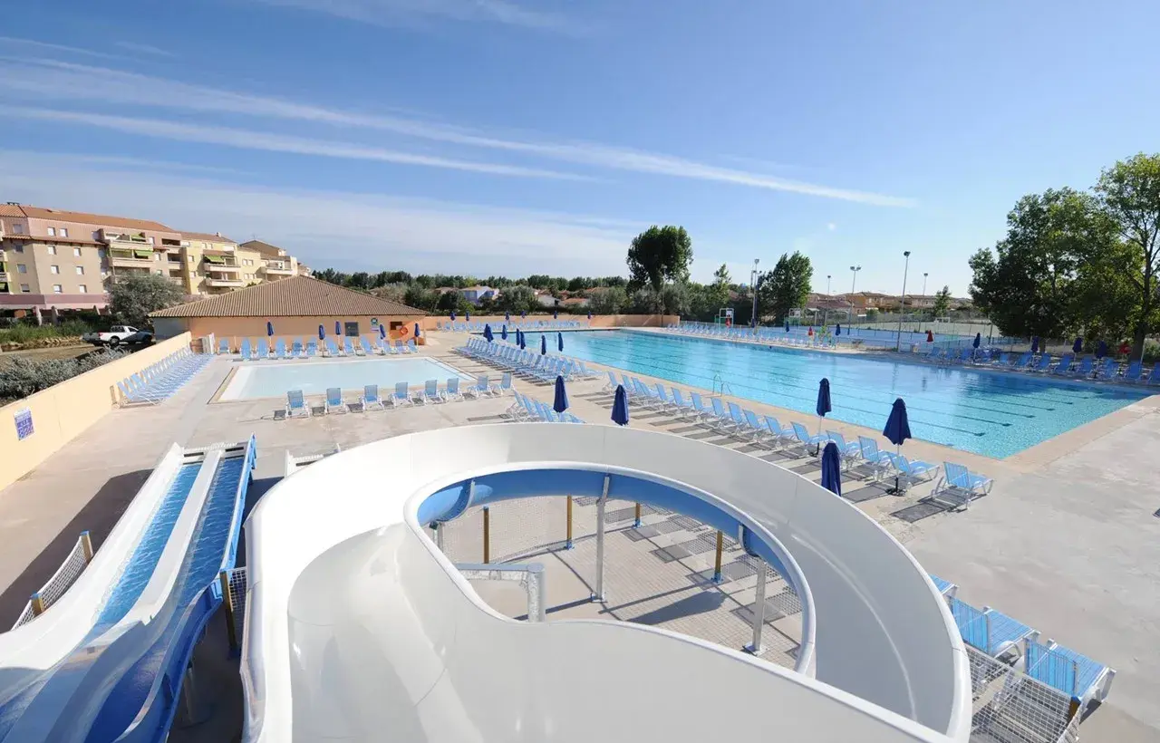 Piscine et toboggans aquatiques dans une aire de camping-car au Grau-du-Roi. Des chaises longues bleues et des parasols sont disposés autour des bassins.