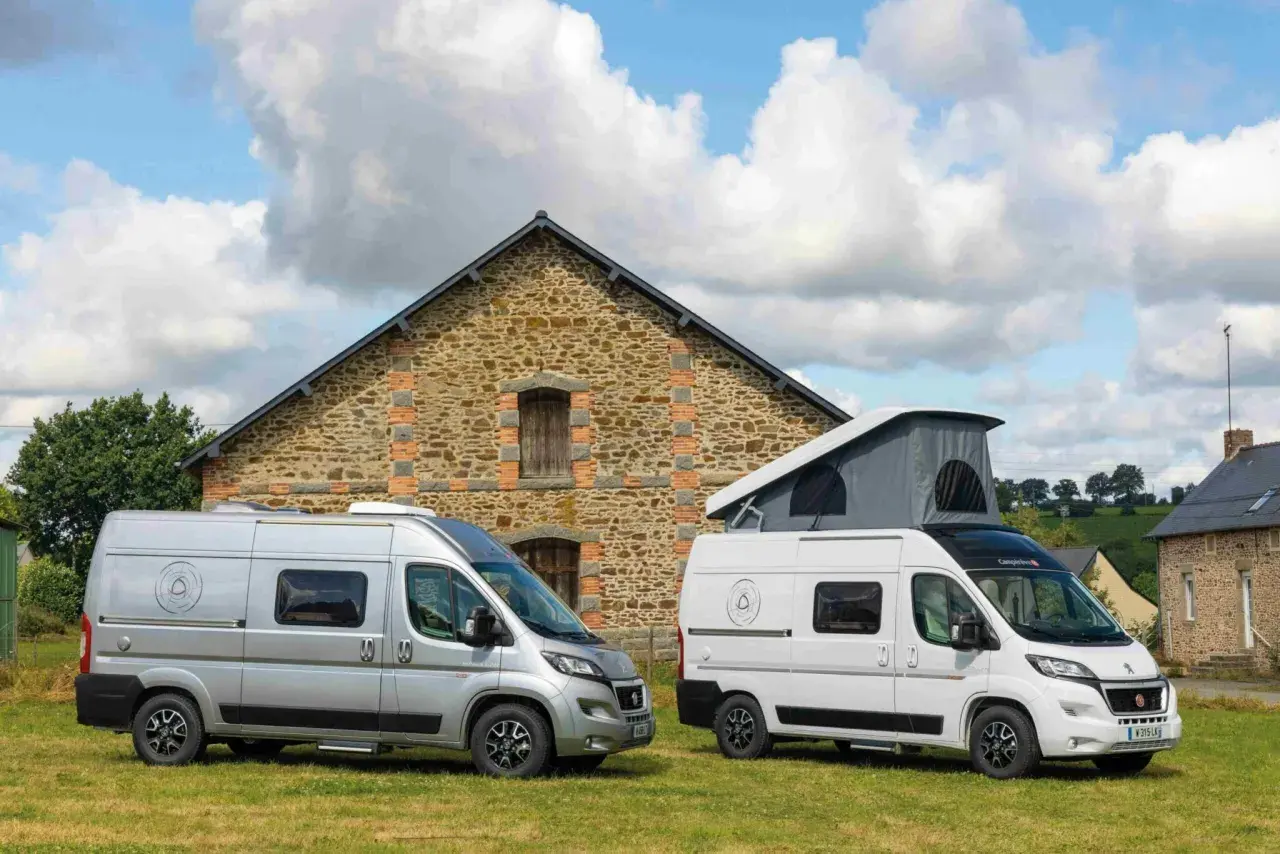 Deux camping-cars, un argenté et un blanc avec toit relevable, devant une bâtisse en pierre. Idéal pour savoir comment choisir son camping-car.