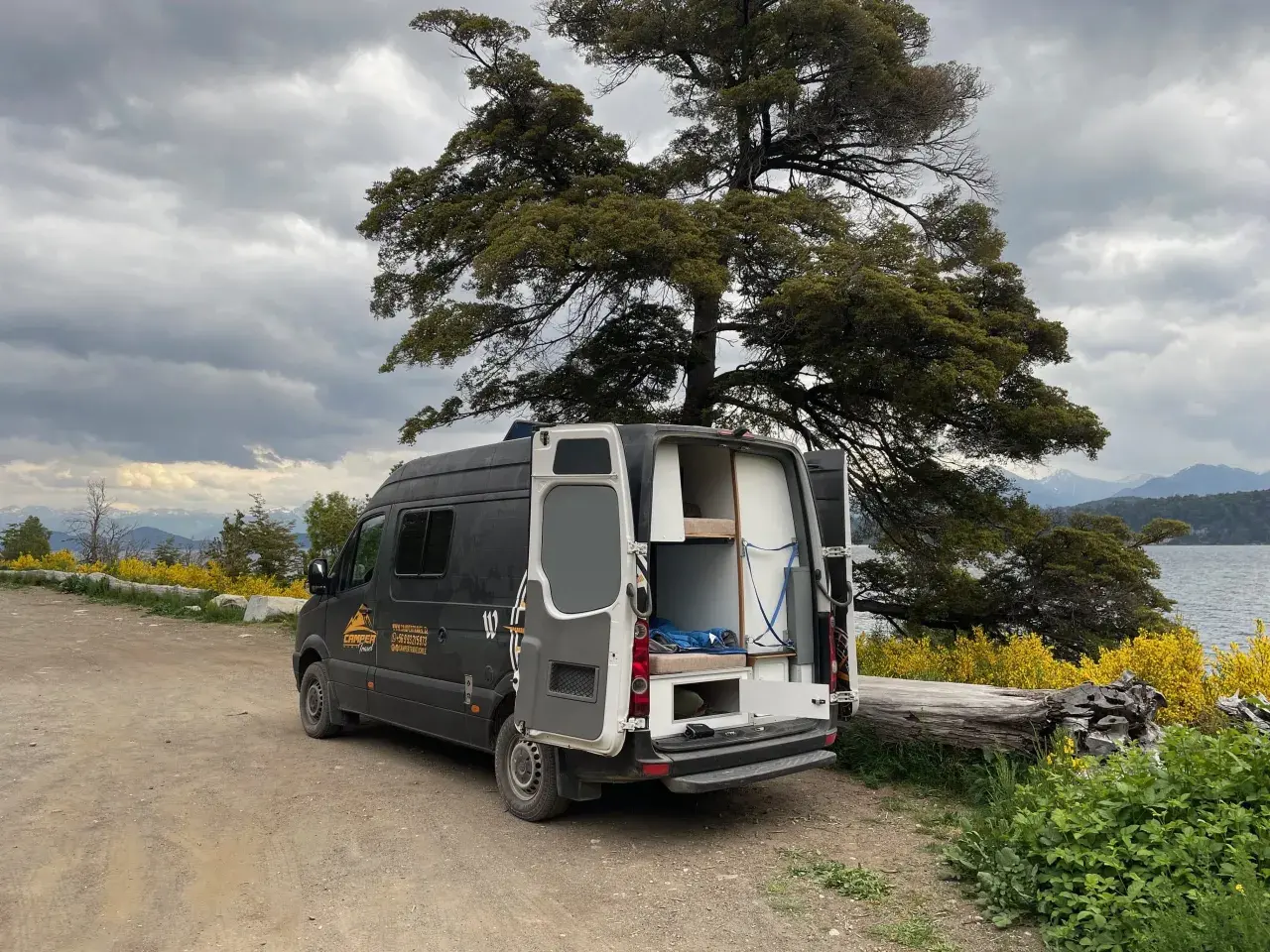 Un camping-car noir avec les portes arrière ouvertes, prêt pour un entretien, est garé près d'un lac sous un ciel nuageux.