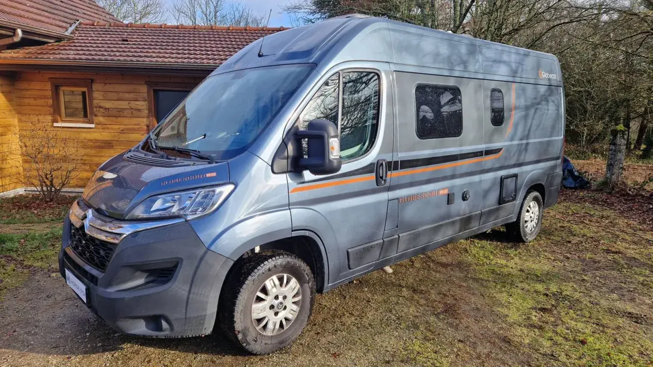 Un camping-car gris, modèle Globecar, est garé devant une maison en bois. Il est prêt pour une aventure avec son look d'Argus.