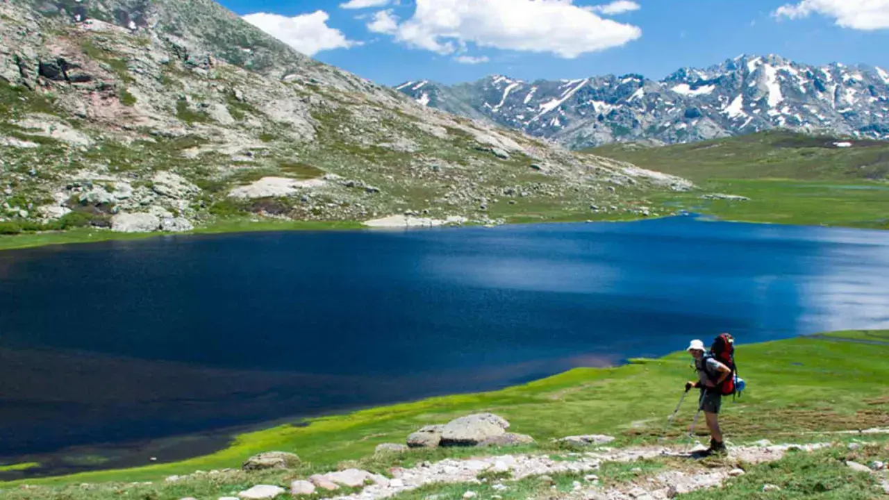 Randonneur sur le GR20 Corse, admirant un lac bleu profond entouré de montagnes verdoyantes et enneigées.