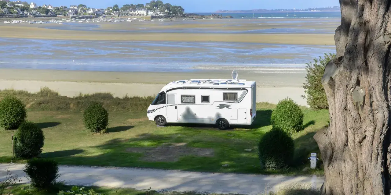 Vue d'une aire de camping-car avec sanitaire, face à une plage et un village côtier. Le camping-car blanc est garé sur l'herbe.