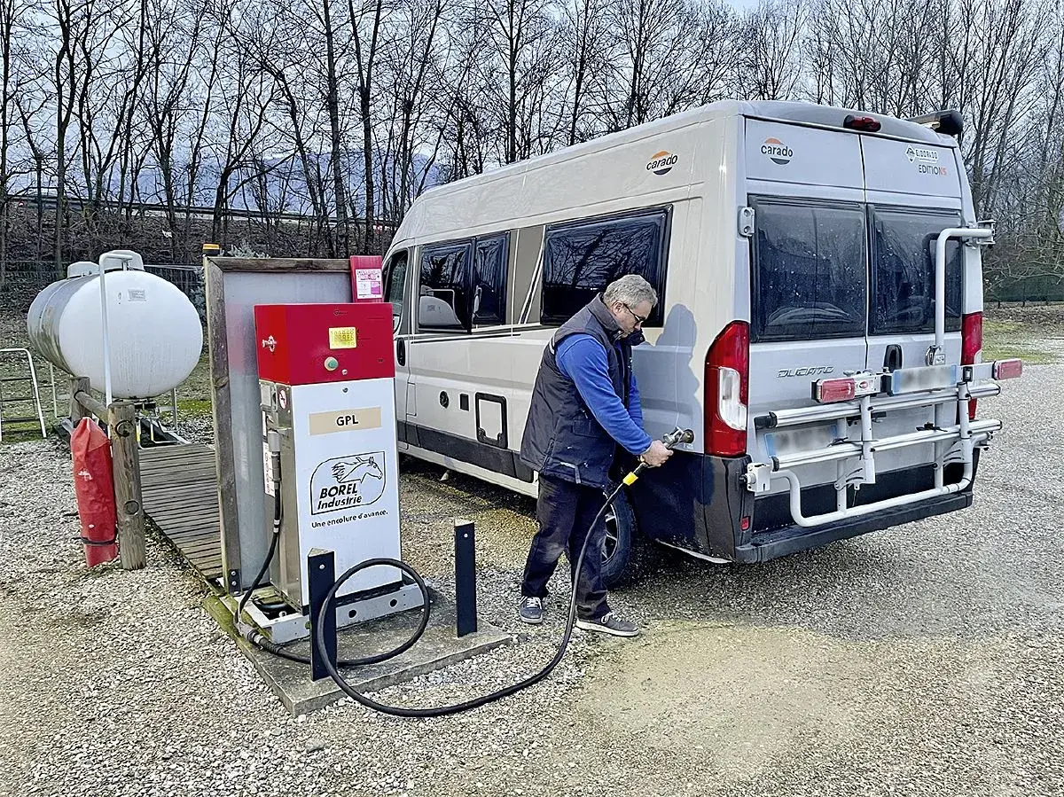 Un homme fait le plein de son camping-car à une installation GPL. Le véhicule est garé à côté d'une pompe à carburant rouge et blanche.