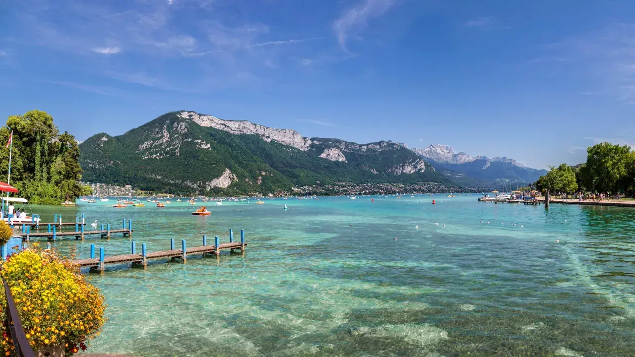 Vue panoramique du lac d'Annecy, avec des montagnes verdoyantes en arrière-plan. Des bateaux et des pédalos animent les eaux turquoise. Un ponton fleuri invite à la promenade.