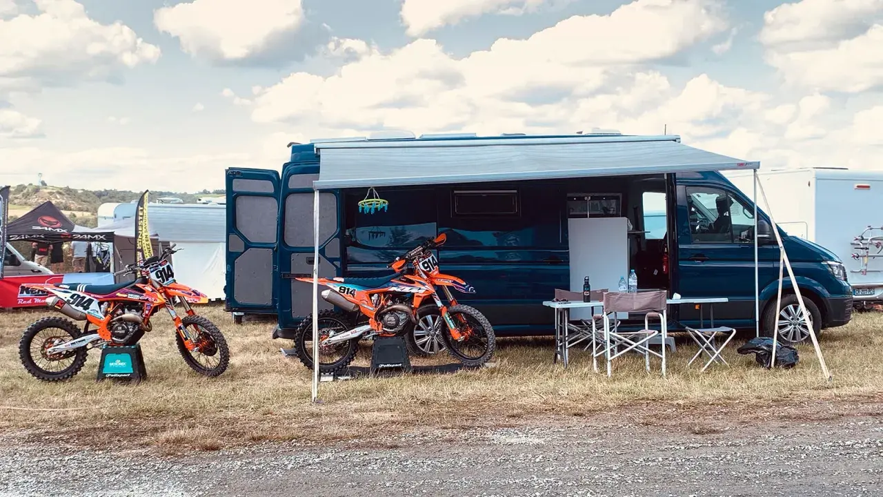 Un fourgon aménagé avec soute moto, prêt pour l'aventure. Deux motos de cross orange attendent devant, sous un ciel nuageux.