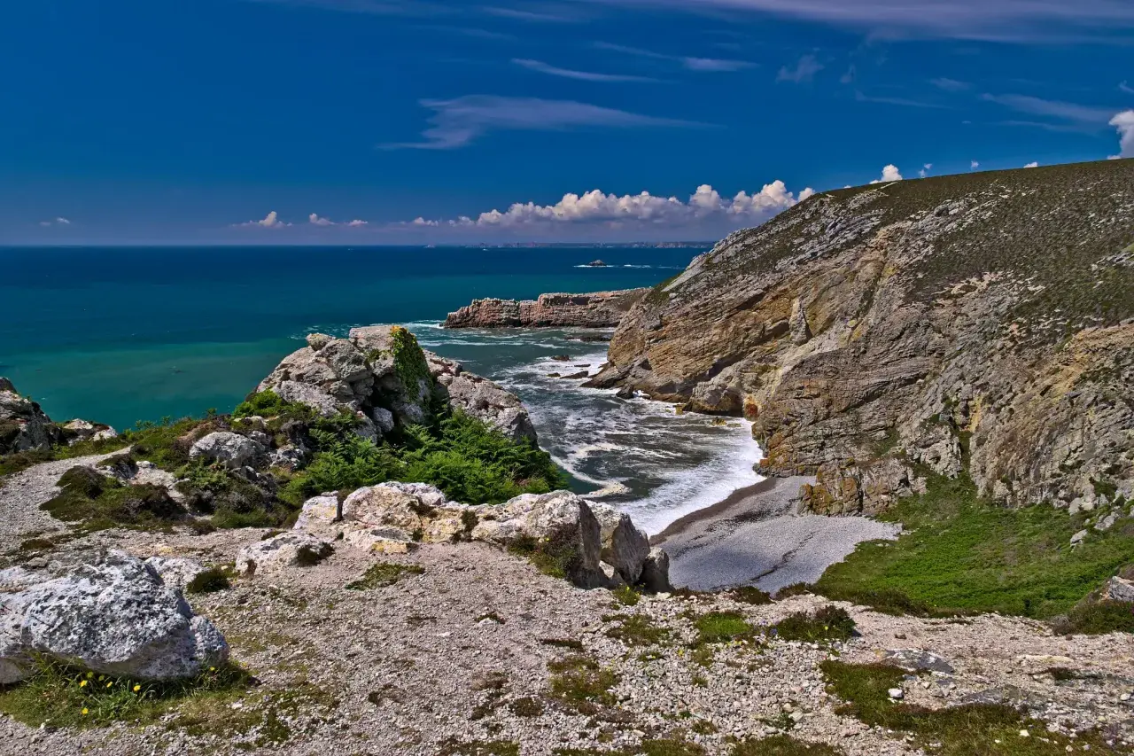 Sentier rocailleux menant au **Cap de la Chèvre**, avec falaises abruptes plongeant dans une mer turquoise et un ciel bleu éclatant.