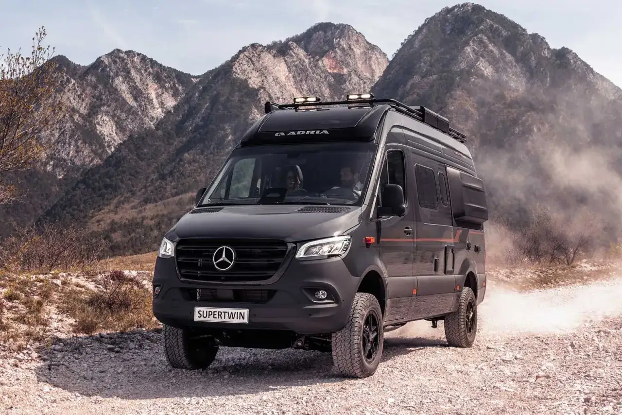 Un camping-car 4x4 Adria Supertwin, prêt pour l'aventure, roule sur un chemin rocailleux avec des montagnes imposantes en arrière-plan.