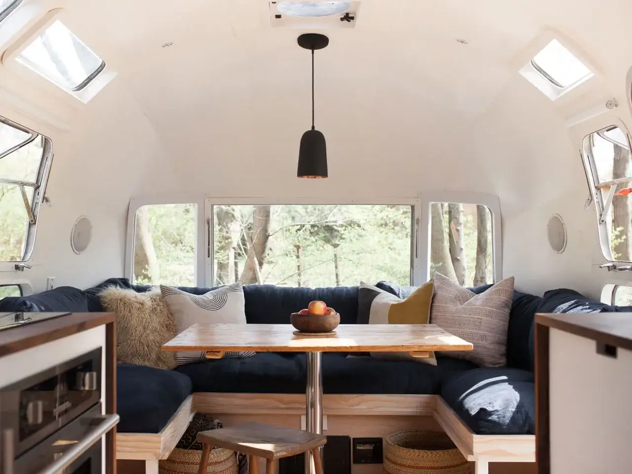 Ambiance cosy dans l'intérieur d'une caravane Airstream. Banquette bleue, table en bois, coussins décoratifs et vue sur la forêt.