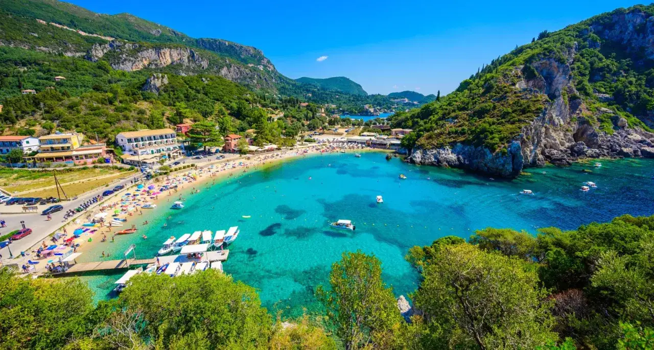 Plage animée à Corfou, eaux turquoise, falaises verdoyantes. Idéal pour savoir que faire à Corfou : baignade, bronzage, bateaux.