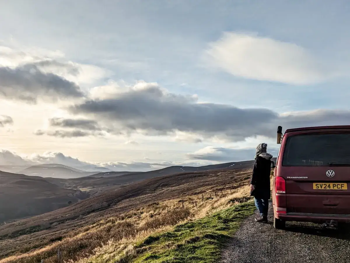 Un voyage en Écosse en camping-car, avec une personne admirant le paysage vallonné sous un ciel nuageux.