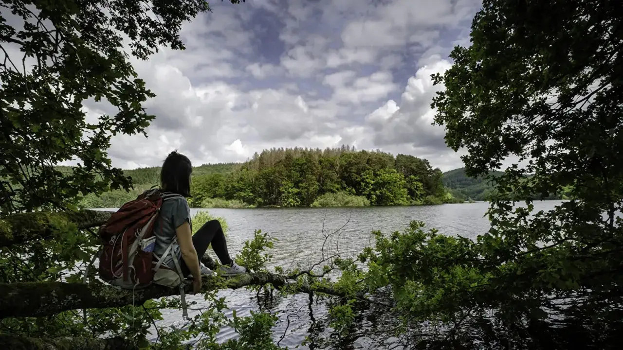 Une randonneuse profite d'une pause au bord d'un lac dans le Morvan, le regard perdu vers la forêt verdoyante.