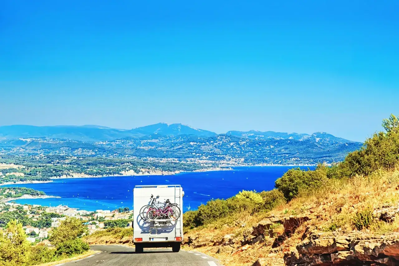 Un camping-car avec v&eacute;los sur un porte-v&eacute;lo, sur une route de campagne avec vue sur une baie bleue et des montagnes. Parfait pour une id&eacute;e sortie week-end camping-car.