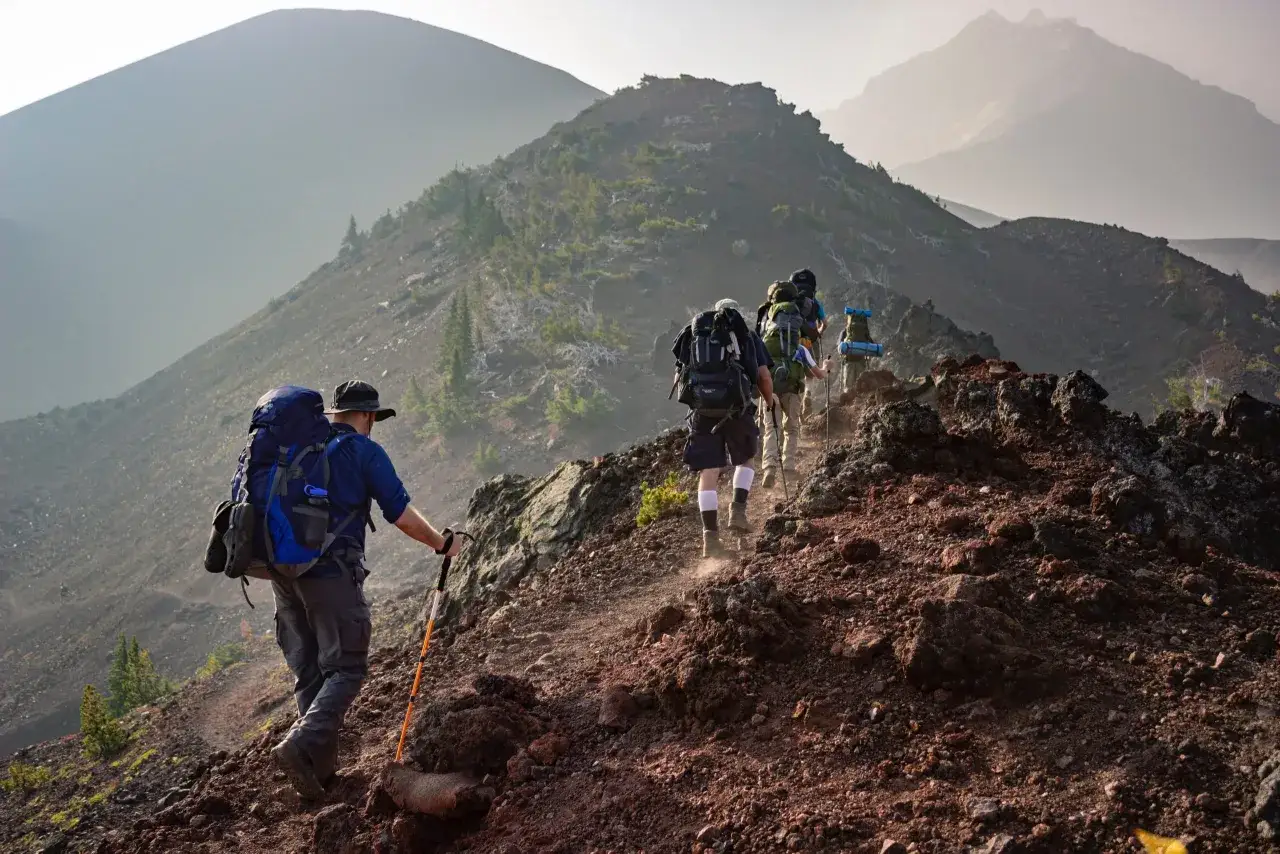 Un groupe de randonneurs progresse sur un sentier rocailleux, sac à dos sur le dos, dans un paysage de montagne brumeux.