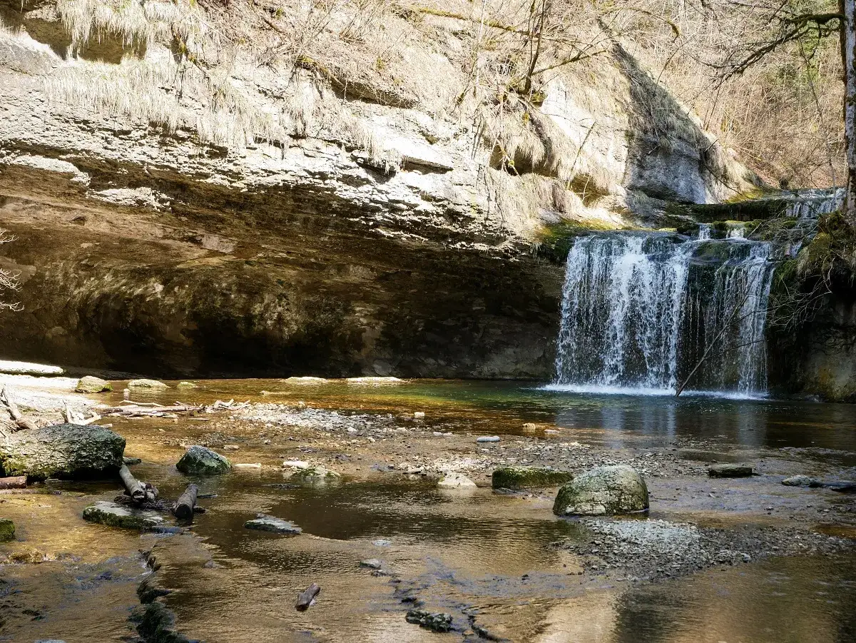 Une belle cascade dans le Jura, avec une grotte rocheuse derrière et une rivière aux eaux claires.