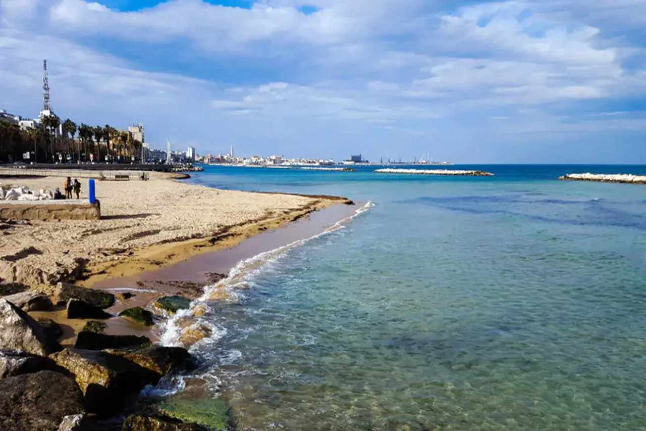 Vue sur la bari plage avec des vagues douces, des rochers et la ville en arri&egrave;re-plan sous un ciel bleu et nuageux.