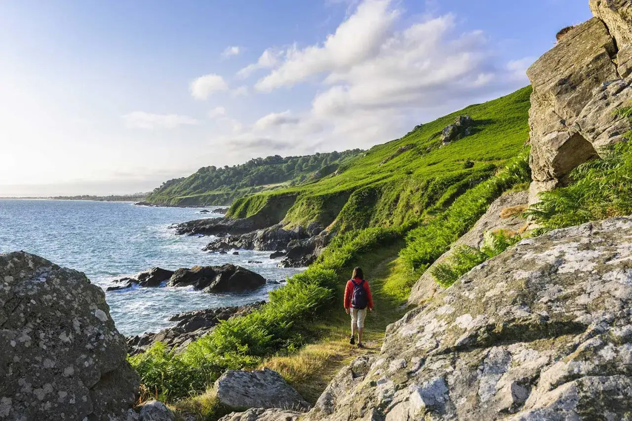 Randonnée sur un sentier côtier verdoyant du GR Cotentin, avec la mer d'un bleu éclatant et des falaises rocheuses.