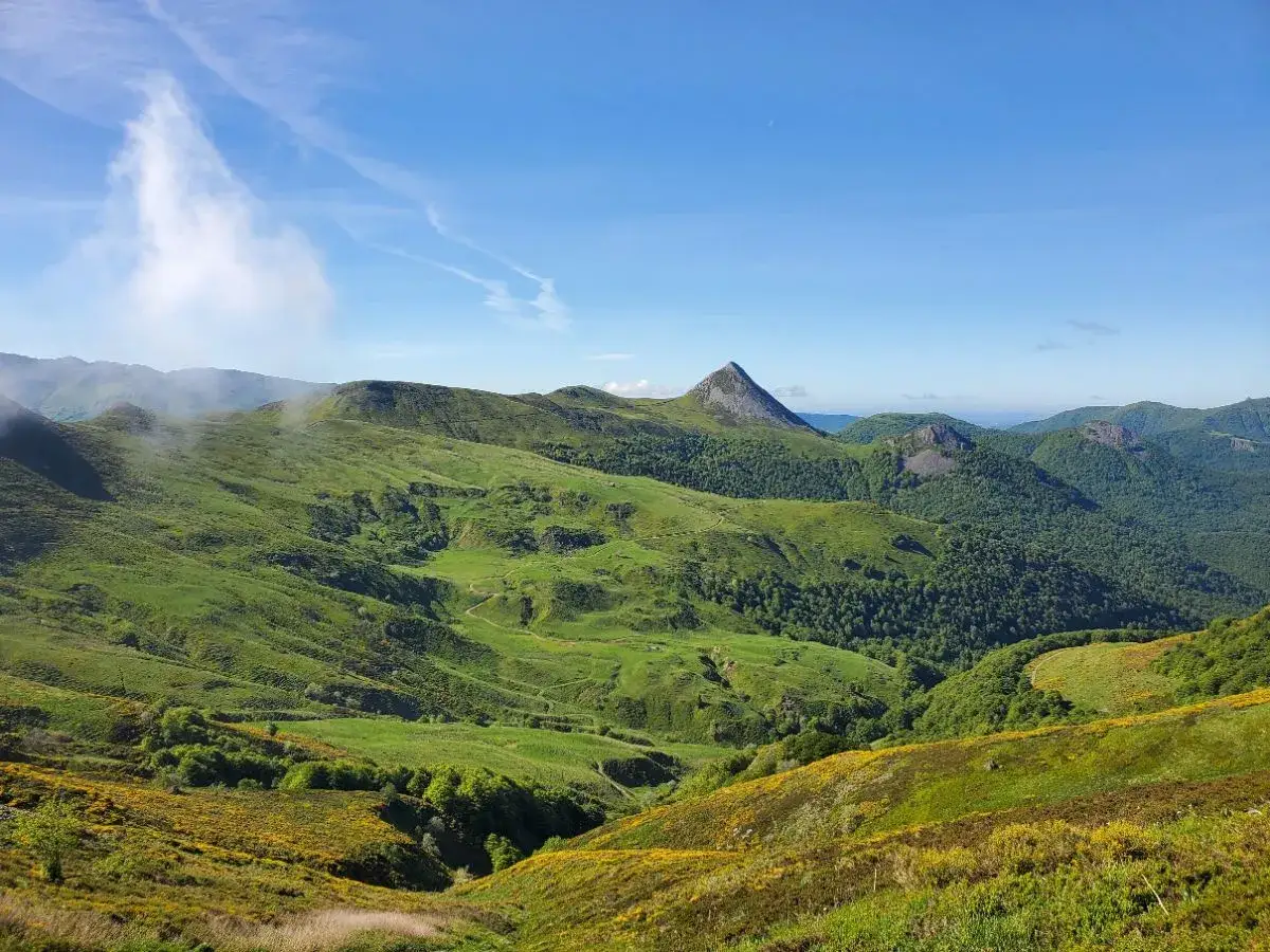 Vue panoramique sur la vallée de la Jordanne, avec ses collines verdoyantes, ses sentiers sinueux et un pic rocheux dominant le paysage sous un ciel bleu.