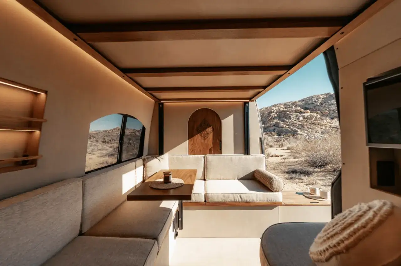 Intérieur d'un camping car petite taille, avec banquettes, table et porte en bois, offrant une vue sur un paysage désertique.