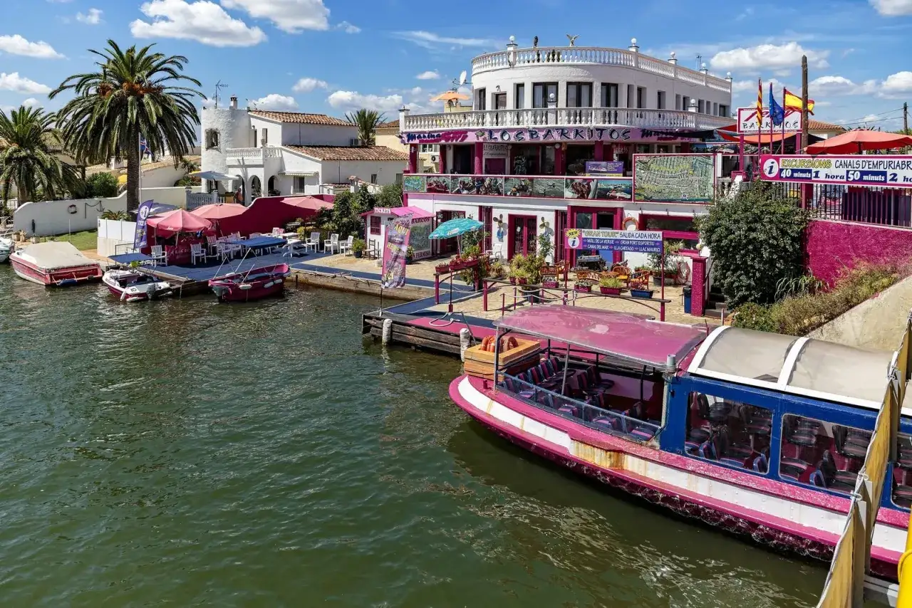 Bateaux roses et blancs amarr&eacute;s le long des canaux d'Empuriabrava, Espagne. Un restaurant color&eacute; avec des parasols roses et des palmiers compl&egrave;te le paysage.