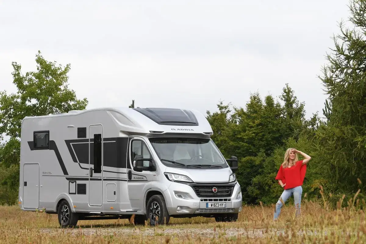 Une femme en tenue décontractée admire un camping-car Adria Matrix 670 SC argenté, prêt pour l'aventure.