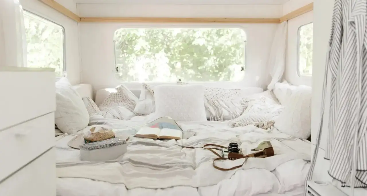 Intérieur d'un camping-car lit pavillon uniquement, décoré de coussins blancs et d'une couverture rayée. Un livre ouvert, un chapeau et un appareil photo vintage invitent au voyage.