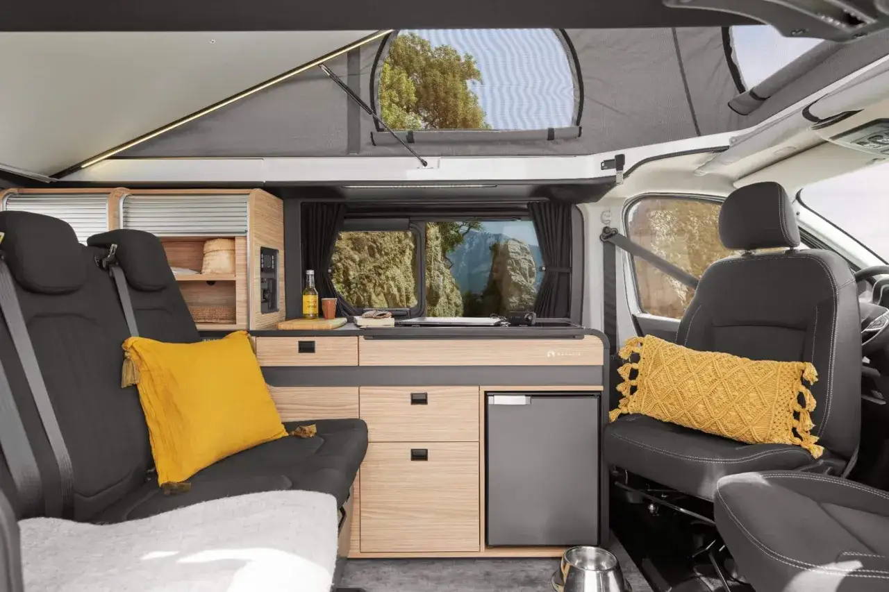 Aménagement intérieur d'un camping-car Bavaria : banquette confortable, cuisine fonctionnelle avec réfrigérateur, et vue panoramique sur la nature.