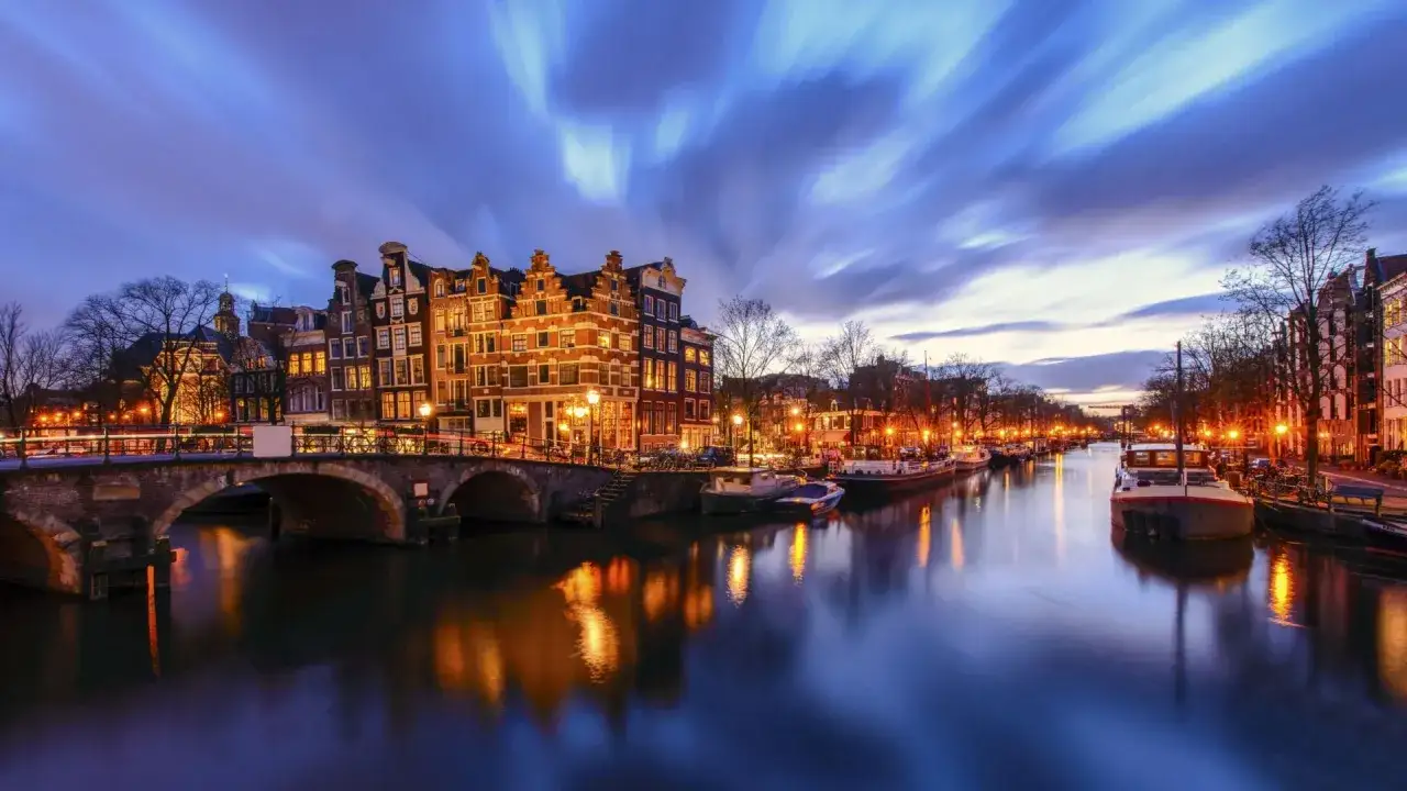 Ambiance nocturne sur un canal d'Amsterdam, avec des maisons illuminées, des bateaux et un pont. Idéal pour découvrir les activités à découvrir.