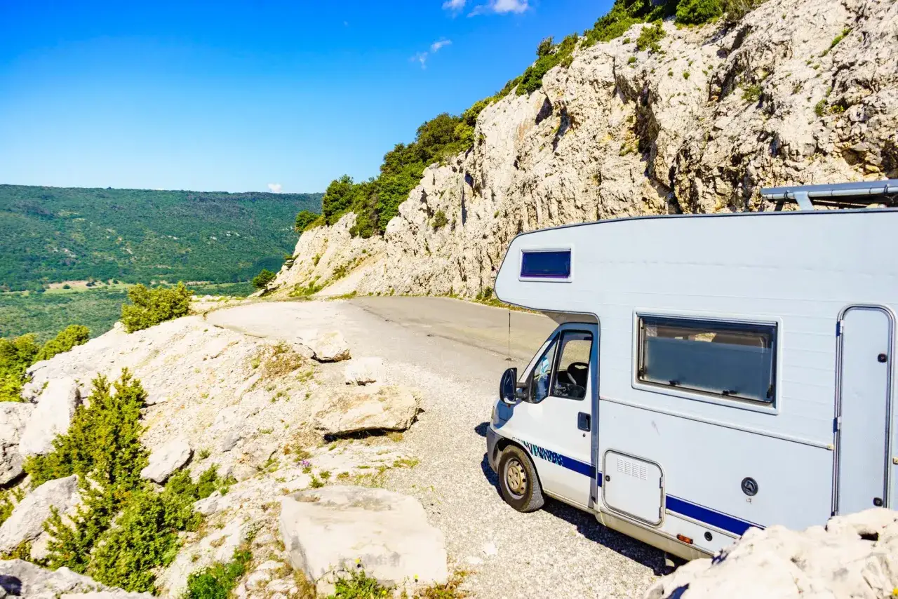 Un camping-car blanc stationné sur une route de montagne rocailleuse, avec une vue panoramique sur une vallée verdoyante.