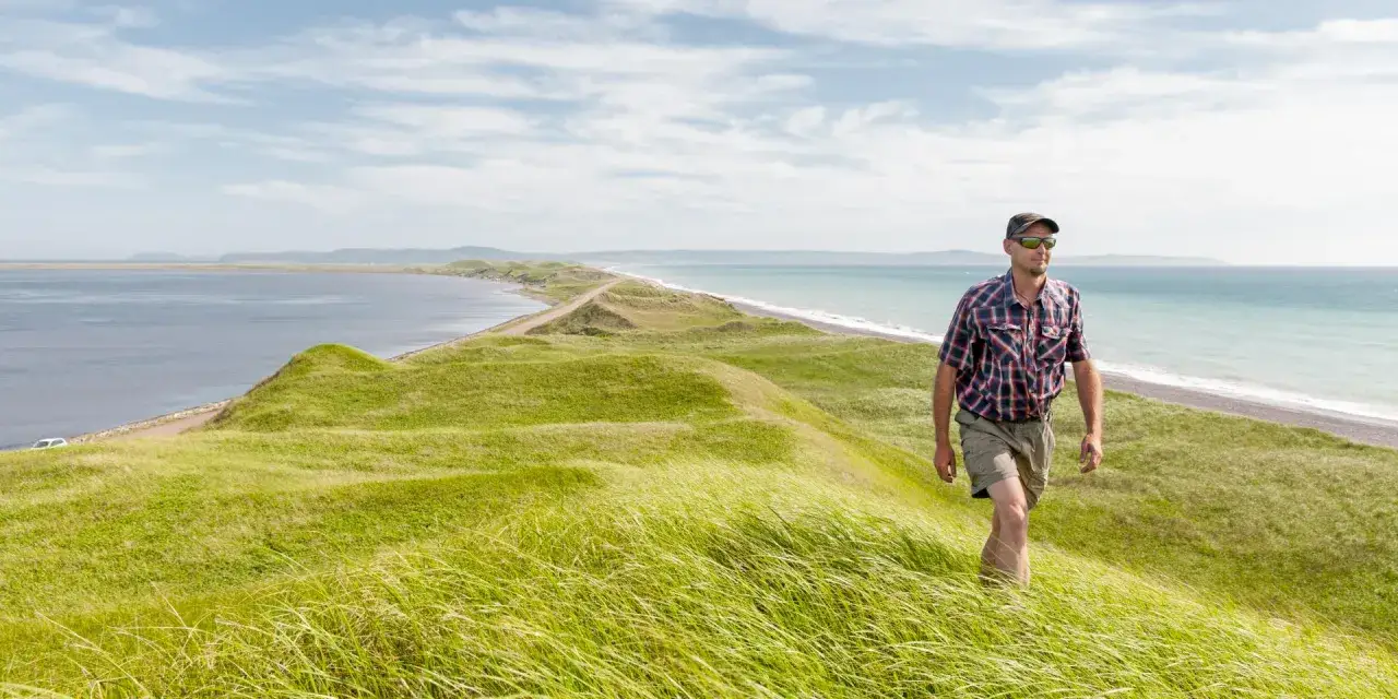 Un homme marche sur une colline verdoyante à Saint-Pierre-et-Miquelon, avec la mer d'un côté et une longue plage de l'autre.