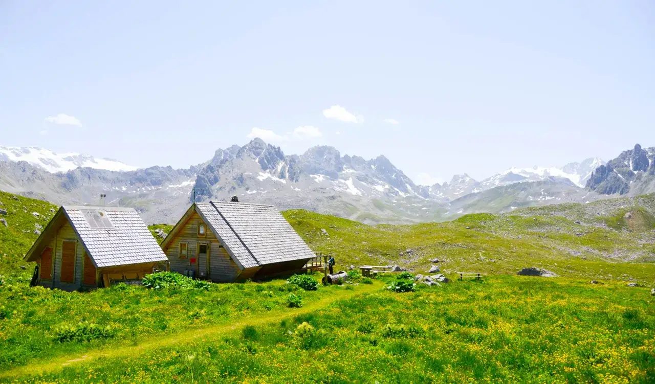 Deux cabanes refuge en bois, nich&eacute;es dans une prairie fleurie, avec des montagnes enneig&eacute;es en arri&egrave;re-plan.