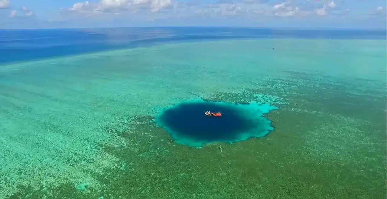 Un bateau rouge et blanc flotte au-dessus du trou du dragon, un ab&icirc;me bleu profond entour&eacute; de r&eacute;cifs coralliens turquoise.
