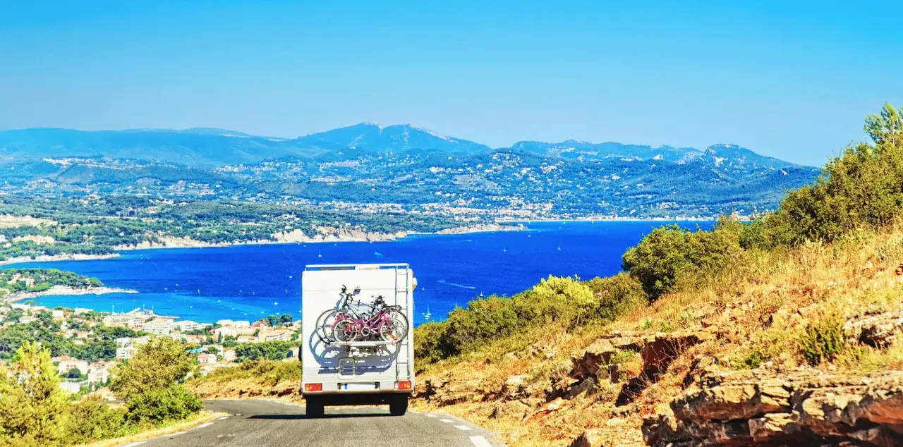 Un camping-car avec v&eacute;los &agrave; l'arri&egrave;re descend une route sinueuse vers une baie bleue scintillante. Un circuit camping-car Italie du Nord promet des vues magnifiques.