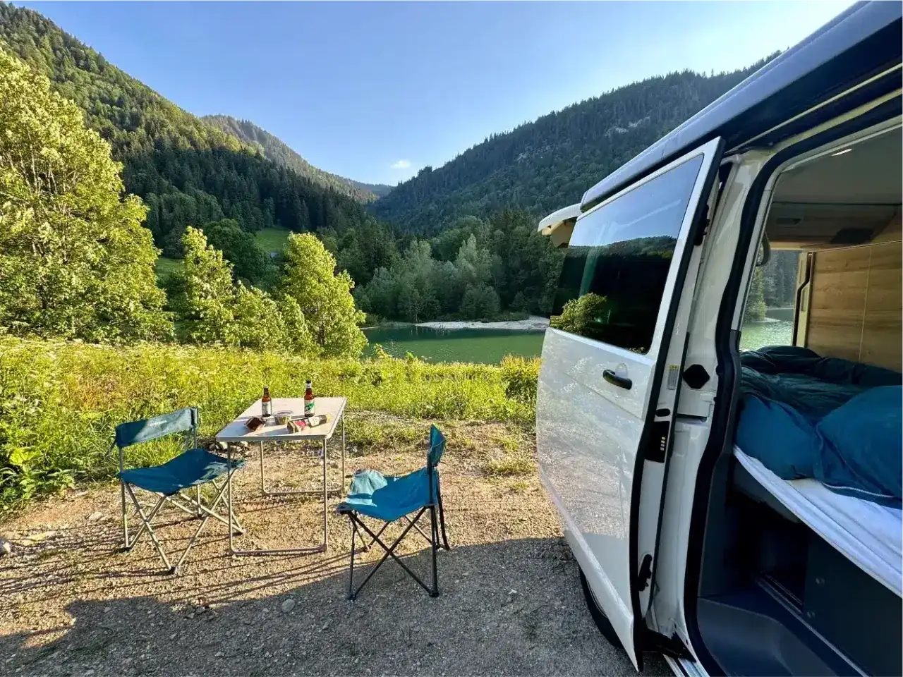 Un coin de paradis pour la Bourgogne en camping-car : table dressée, chaises bleues, lac paisible et montagnes verdoyantes.