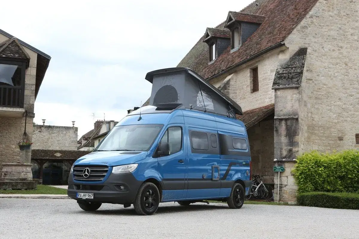 Un porteur camping-car bleu avec toit relevable est garé devant une vieille bâtisse en pierre.