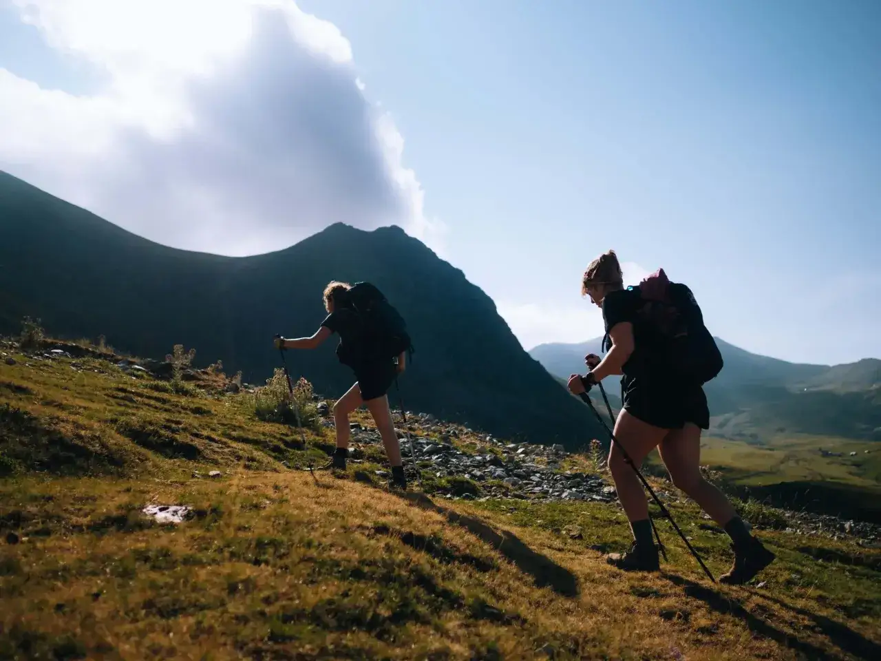 Deux randonneuses gravissent une pente herbeuse, bâtons à la main, sous un ciel bleu éclatant. La montagne s'étend derrière elles.