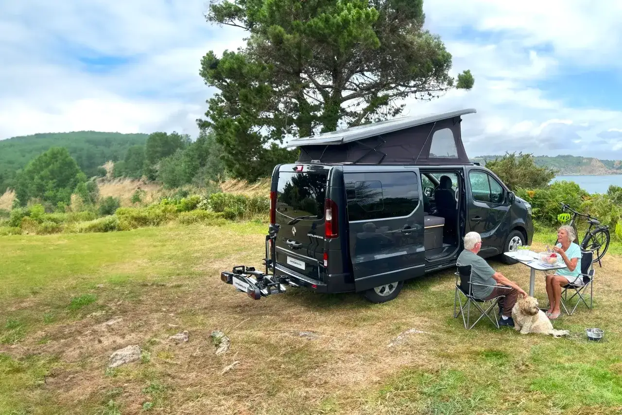 Un couple et leur chien profitent d'un repas devant leur camping-car aménagé, avec une vue sur la mer et la campagne.