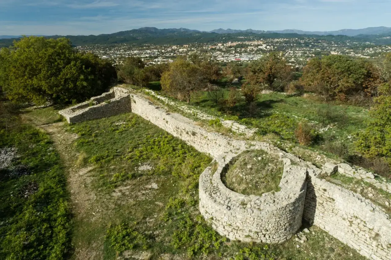Vestiges de l'oppidum de Jastres, murs de pierre anciens sur une colline verdoyante, avec une ville et des montagnes en arri&egrave;re-plan.